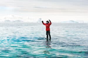 Person in a red parka stands in icy water with glaciers and icebergs in the distance, arms raised in triumph.