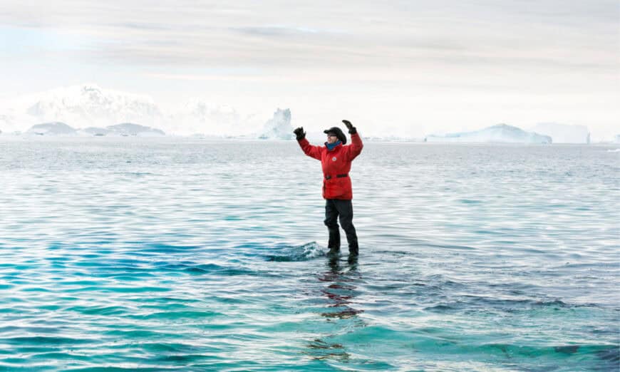 STanding-on-water | Photography and video news, reviews and tips | Henry's Camera Person in a red parka stands in icy water with glaciers and icebergs in the distance, arms raised in triumph.