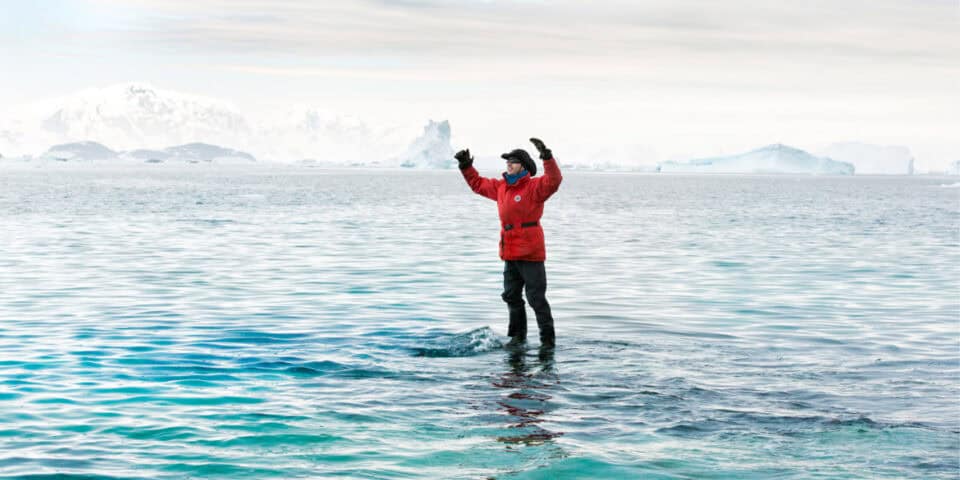 Person in a red parka stands in icy water with glaciers and icebergs in the distance, arms raised in triumph.