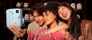 Three friends posing for a selfie in a bowling alley, with shelves of white bowling shoes behind them and wearing colorful outfits.