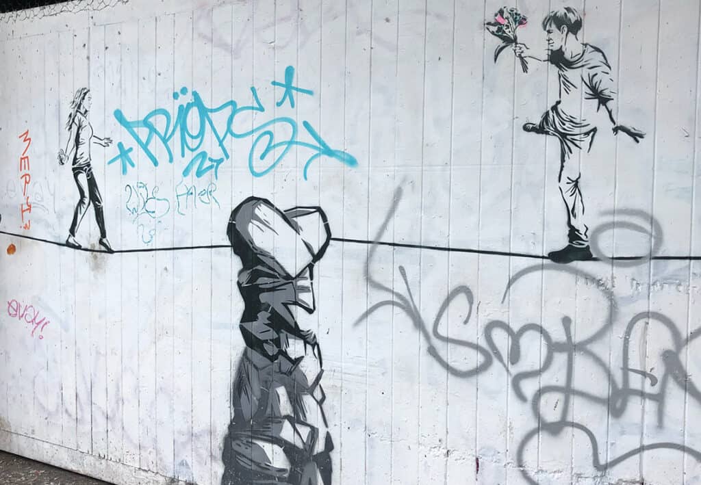 Street-art mural of a man on the right holding flowers toward a woman walking away on the left, with a large rock-like figure in the foreground and blue graffiti tags in the background.