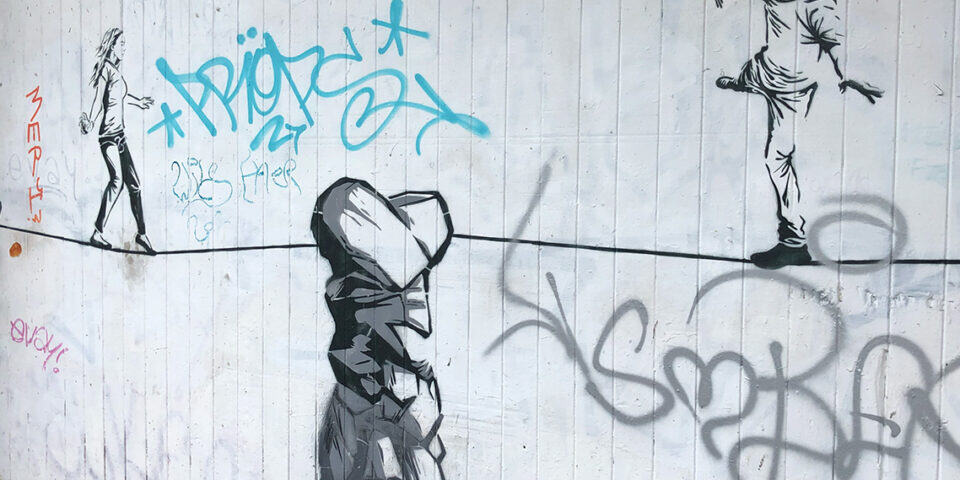 Street-art mural of a man on the right holding flowers toward a woman walking away on the left, with a large rock-like figure in the foreground and blue graffiti tags in the background.