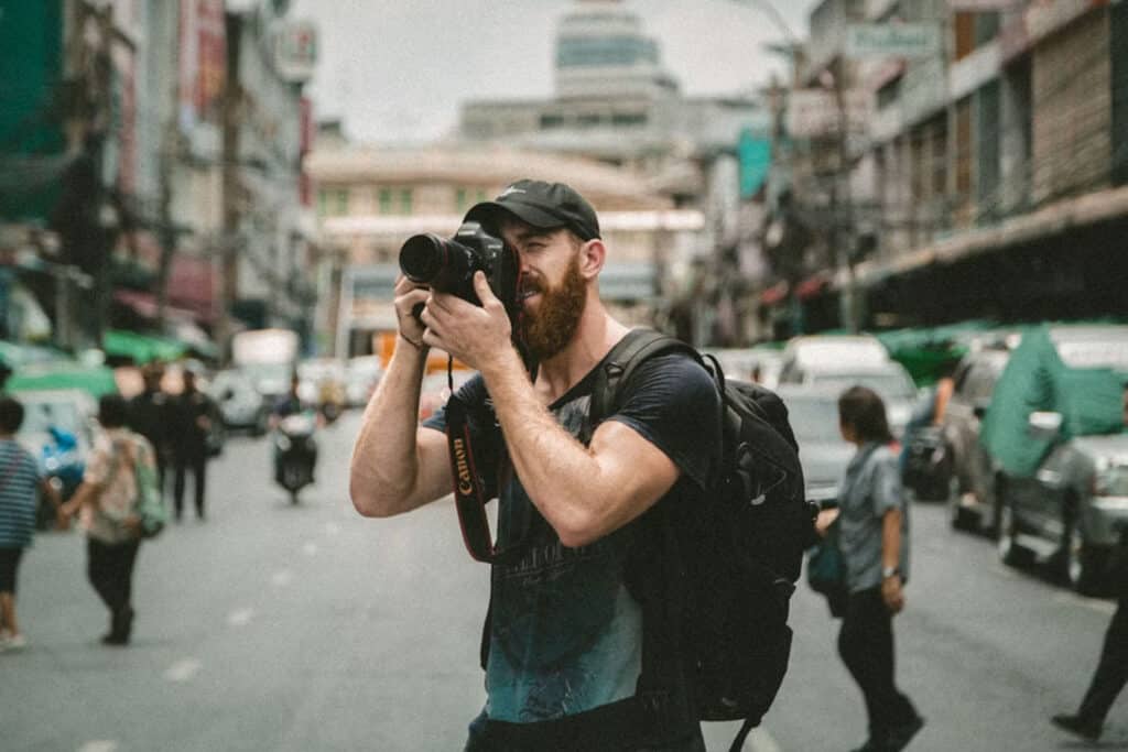 Bearded man in a black cap and backpack uses a DSLR to photograph a busy city street.