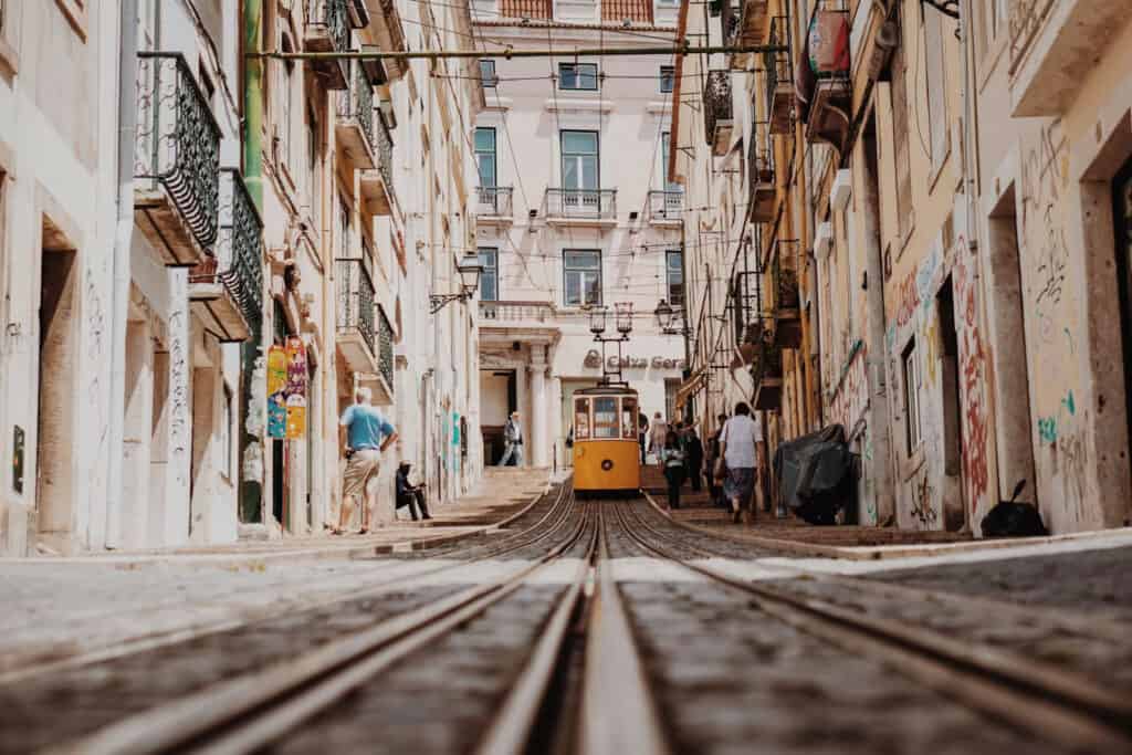 Yellow tram on a narrow city street with old buildings, pedestrians, and converging tram tracks in the foreground.