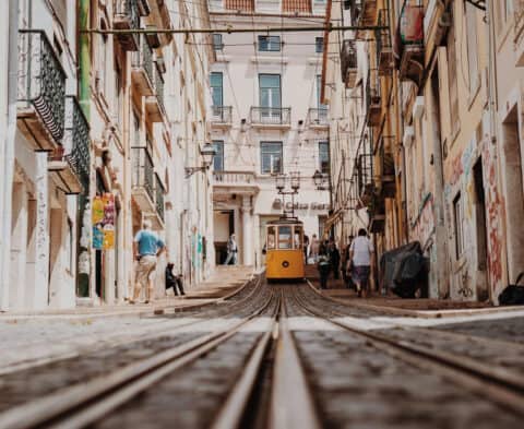 Yellow tram on a narrow city street with old buildings, pedestrians, and converging tram tracks in the foreground.