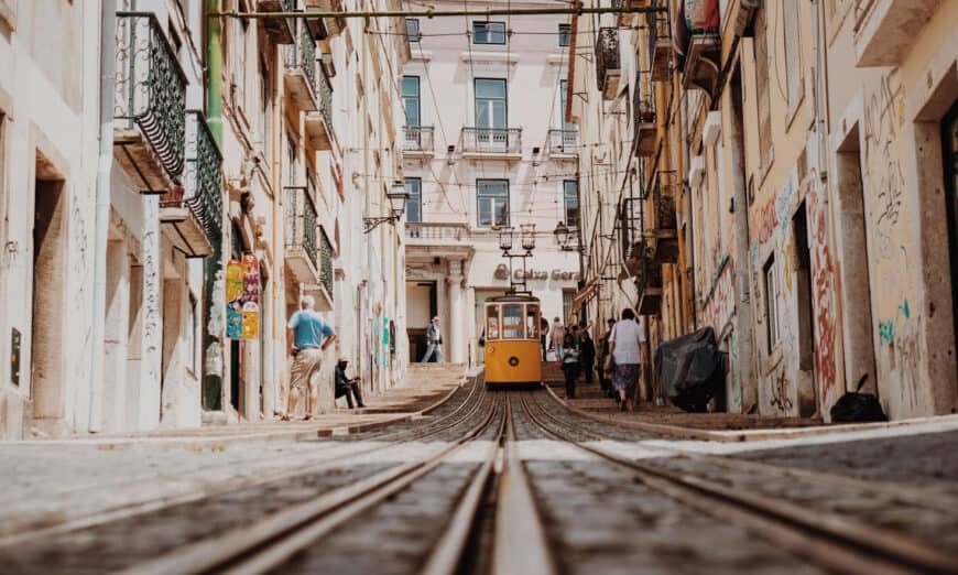 Yellow tram on a narrow city street with old buildings, pedestrians, and converging tram tracks in the foreground.