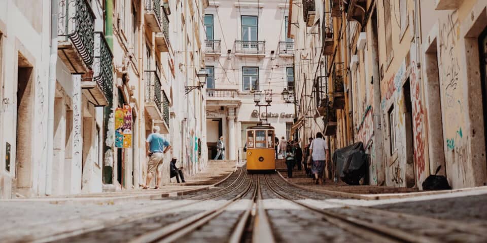 Yellow tram on a narrow city street with old buildings, pedestrians, and converging tram tracks in the foreground.
