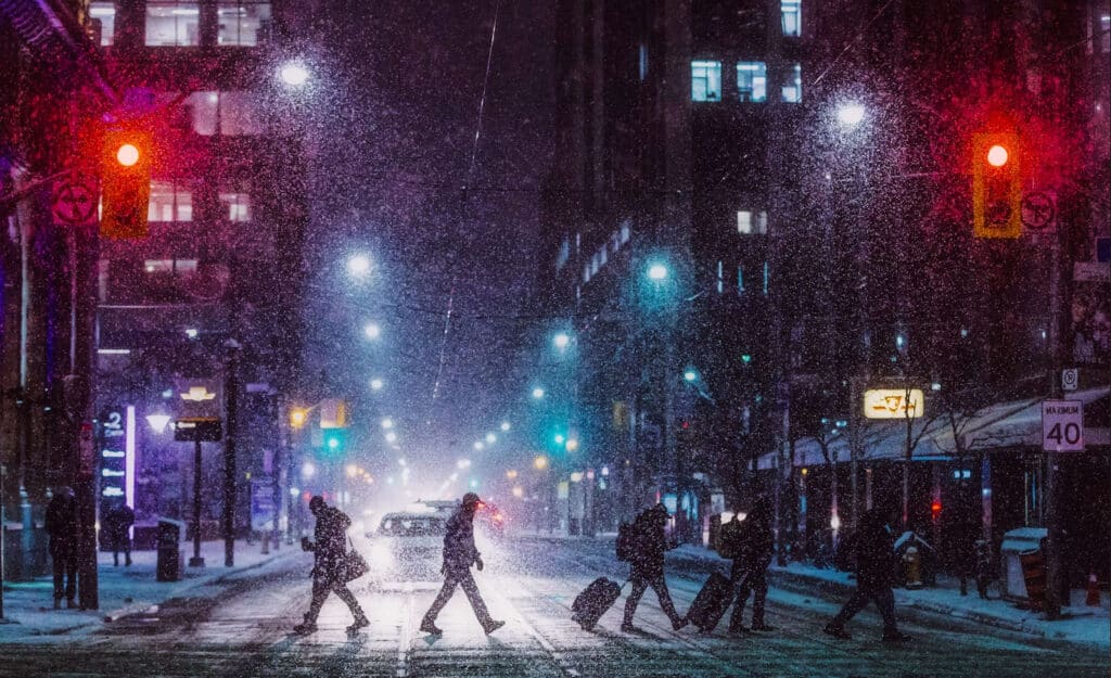 Nighttime city street in heavy snowfall with pedestrians crossing a lit crosswalk and a car in the distance.