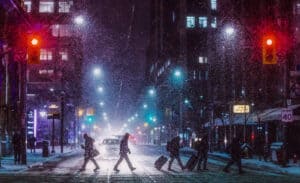 Nighttime city street in heavy snowfall with pedestrians crossing a lit crosswalk and a car in the distance.