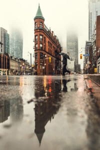 Toronto-Street | Photography and video news, reviews and tips | Henry's Camera Rainy city street with a person under an umbrella; a turreted red brick building and tall skyscrapers in the misty background, reflected in a puddle.