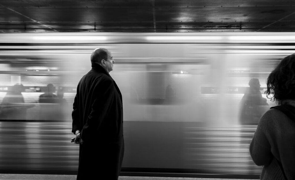 Man waiting on a subway platform as a fast-moving train blurs past in the background, with a person on the right.