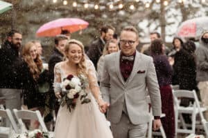 Bride and groom walk hand in hand down the aisle in snowfall, bride holding a white-pink bouquet, guests smile in the background.