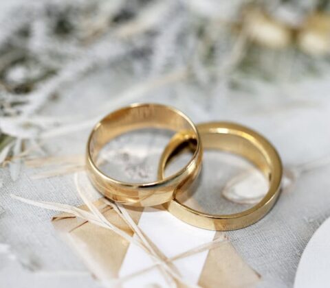 Two gold wedding bands resting on a lacey, pale backdrop with ribbons and greenery in soft focus to convey a wedding theme.