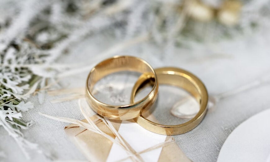 Wedding | Photography and video news, reviews and tips | Henry's Camera Two gold wedding bands resting on a lacey, pale backdrop with ribbons and greenery in soft focus to convey a wedding theme.