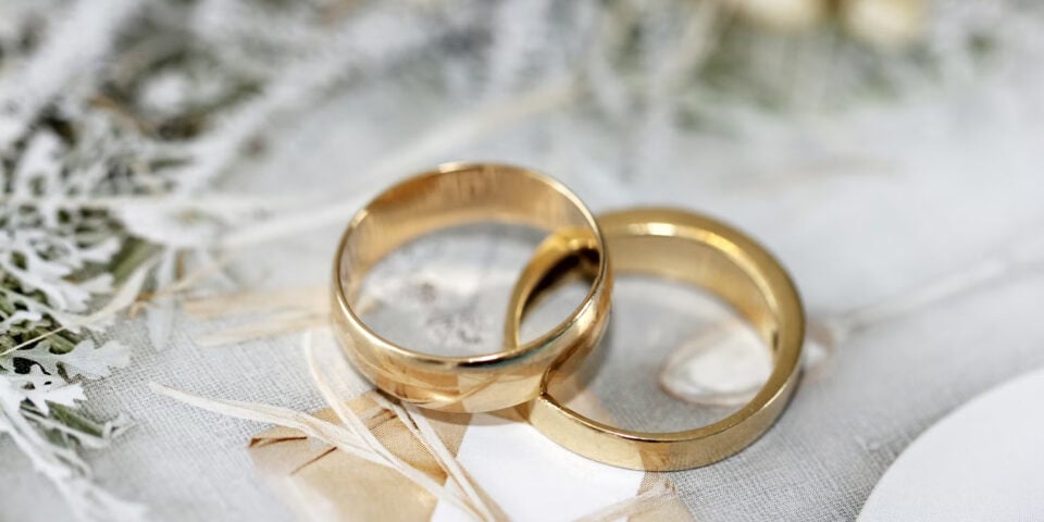 Two gold wedding bands resting on a lacey, pale backdrop with ribbons and greenery in soft focus to convey a wedding theme.