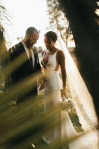 Bride in a white gown and groom in a dark suit stand together in a sunlit garden at golden hour, veil flowing and bouquet in hand.