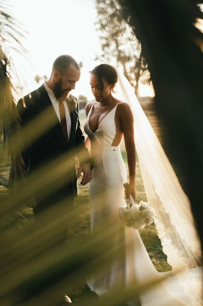 Bride in a white gown and groom in a dark suit stand together in a sunlit garden at golden hour, veil flowing and bouquet in hand.
