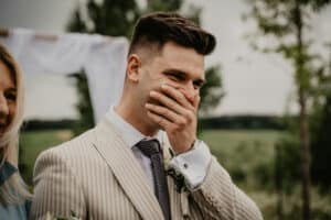 Man in a beige pinstripe suit covers his mouth while laughing at an outdoor wedding venue.