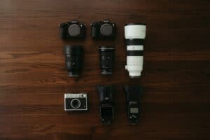 Flat lay of photography gear on a wooden table: two Sony mirrorless bodies, several lenses, a large white telephoto lens, a compact camera, and two studio flashes.