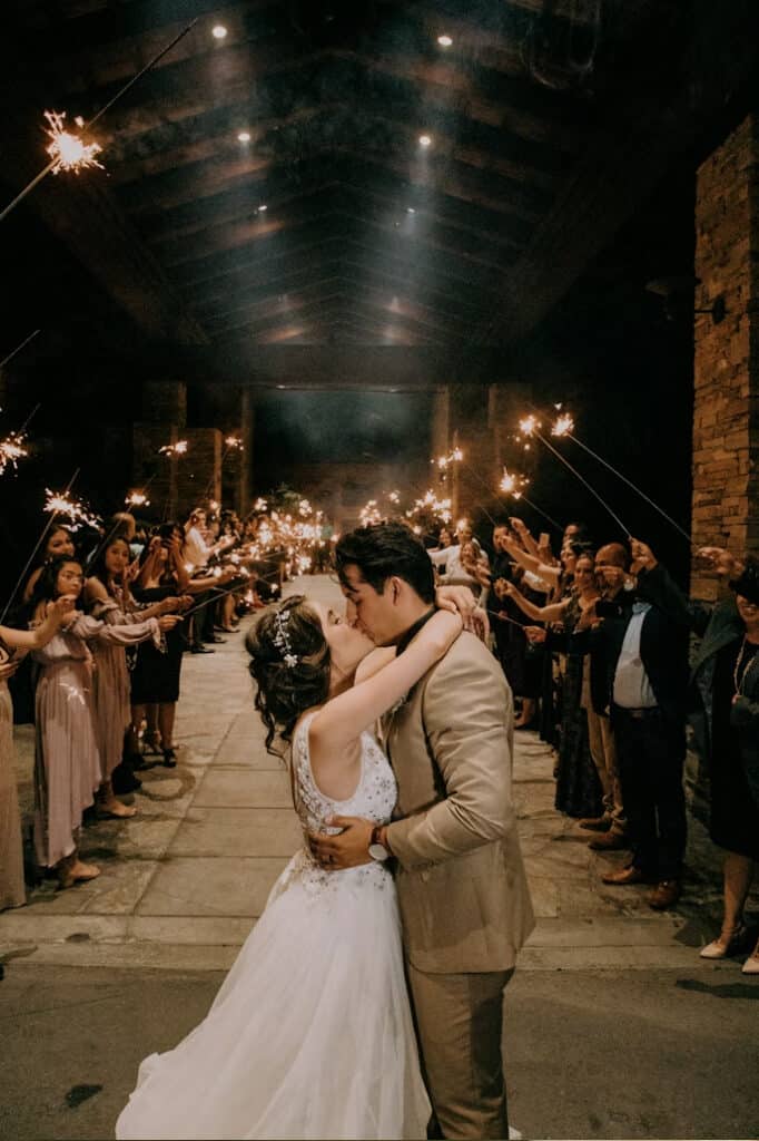 Bride and groom kiss in the center aisle as guests hold sparklers along a stone venue corridor.
