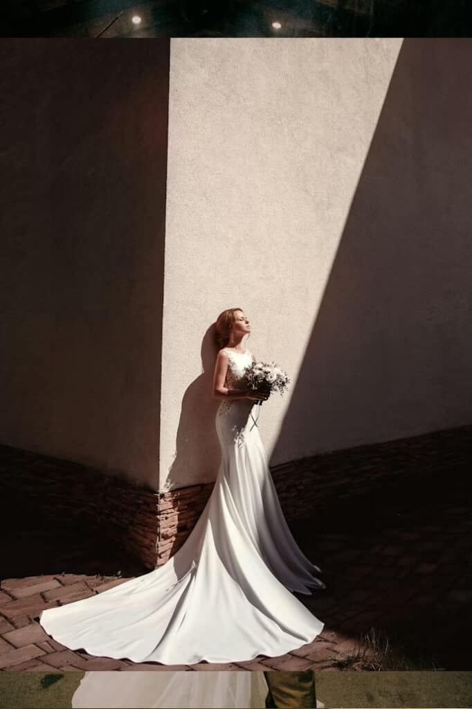 Bride in a long white wedding gown holding a bouquet, leaning against a sunlit wall with a dramatic diagonal shadow.