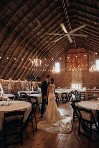 Bride and groom share a kiss on a wooden barn dance floor, surrounded by round tables and warm string lights.