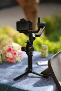 Camera mounted on a handheld three-axis gimbal on a table outdoors, with a bouquet and can in the background.