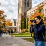 Woman with a backpack shoots with a camera on a university campus as autumn leaves fall. A photo-friendly alt text should be concise; this version mentions subject, action, setting, and season without extra details.